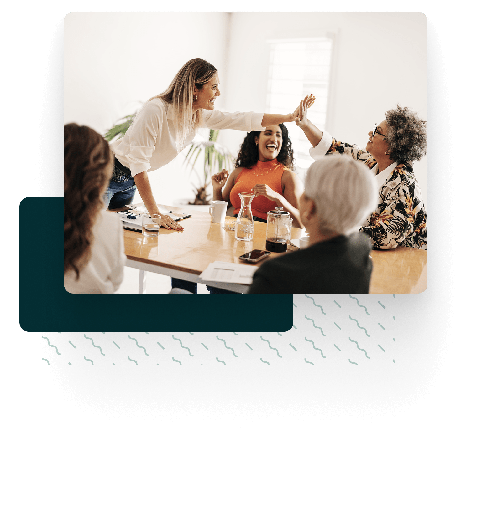 Quatre personnes sont assises autour d'une table pendant une réunion ; deux femmes, souriantes, se penchent de l'autre côté de la table pour se congratuler, tandis que les autres regardent et sourient.