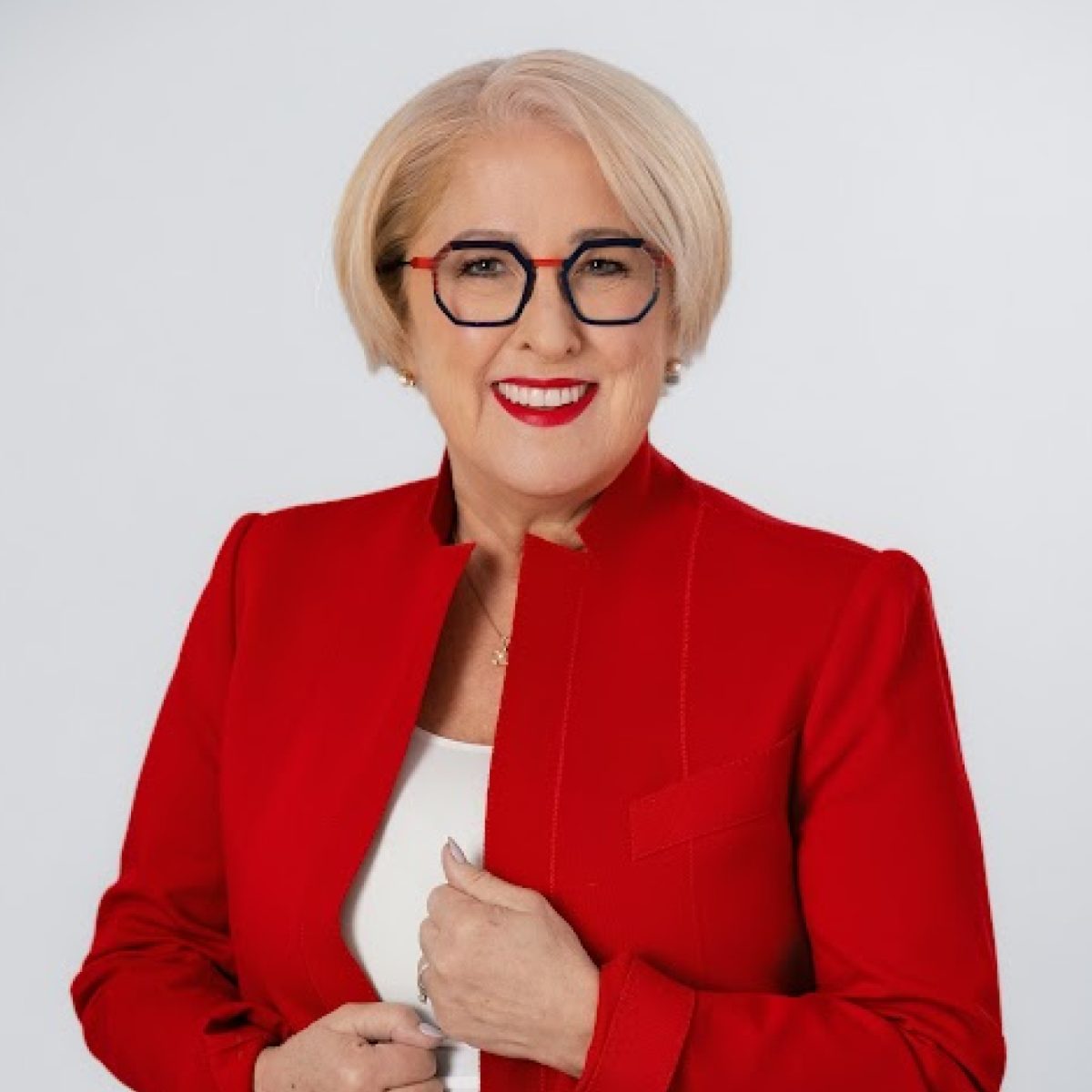 photo_ruth Femme souriante aux cheveux blonds courts, portant des lunettes à monture rouge et du rouge à lèvres rouge vif, vêtue d'un blazer rouge par-dessus un haut blanc, debout devant un fond clair et uni.
